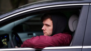 Kırmızı ceketli, siyah kapüşonlu, traşsız genç bir adam arabanın direksiyonunda oturup pencereden dışarı bakıyor.