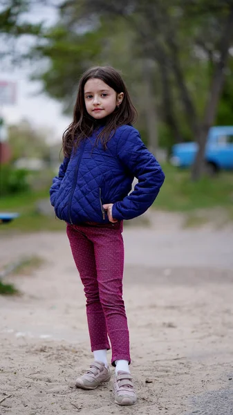 一个长着蓬松长发 穿着蓝色夹克的漂亮小女孩在操场上的一个公园里摆姿势的画像 从侧面看孩子们笑得很好 — 图库照片