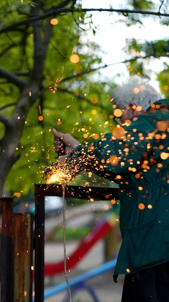 Craftsman in glasses cutting metal with disk grinder outdoors with blurred sparks.