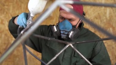 Mavi koruyucu maskeli ve yeşil ceketli bir işçi havasız spreyli metal ürünleri boyuyor. Yavaş çekim