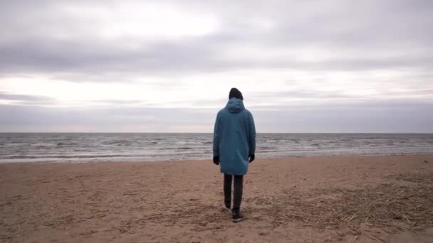 Flicka klädd i en blå regnrock och promenader längs stranden i ett kallt hav — Stockvideo