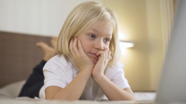 Un mignon garçon d'âge préscolaire couché sur le lit et regardant une vidéo sur un ordinateur portable et riant. Enseignement à distance en ligne . — Video