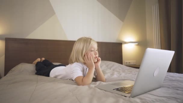 Egy aranyos óvodás fiú fekszik az ágyon és egy videót néz egy laptopon. Távolsági tanulás Online oktatás. — Stock videók