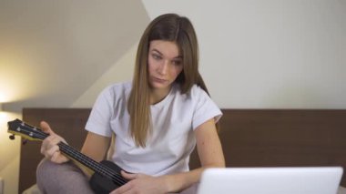 Ukulele çalmayı dizüstü bilgisayarla öğrenen genç bir kadın. Çevrimiçi Eğitimde Uzaklık Öğrenme