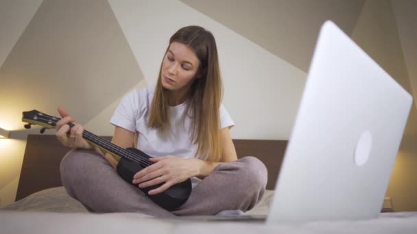 Mujer linda joven aprendiendo a tocar el ukelele usando el ordenador portátil en casa. Educación a distancia Educación en línea — Vídeos de Stock