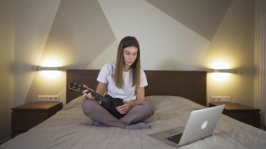 Bilgisayarla ukulele oynayan genç bir kadın, karantinada online kursla oynamayı öğreniyor. Çevrimiçi Eğitimde Uzaklık Öğrenme