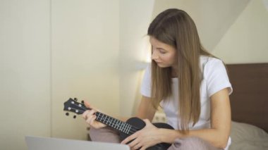 Genç bir kadın dizüstü bilgisayar monitöründen ukulele çalmayı öğreniyor, karantinada evde online öğretim programı izliyor. Çevrimiçi Eğitimde Uzaklık Öğrenme