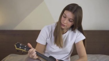 Yatakta Ukulele çalan ve karantinada dizüstü bilgisayar kullanan neşeli kadın. Çevrimiçi Eğitimde Uzaklık Öğrenme