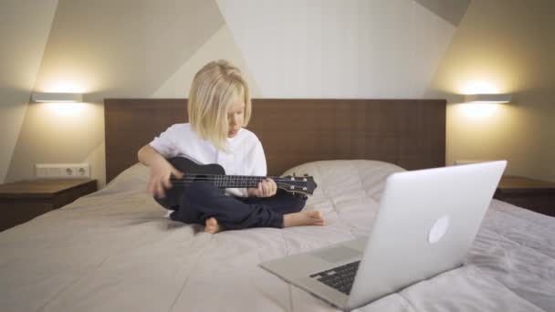 Educación a distancia en línea. Un niño de preescolar aprendiendo a tocar el ukelele y cantando en un portátil en casa . — Vídeos de Stock