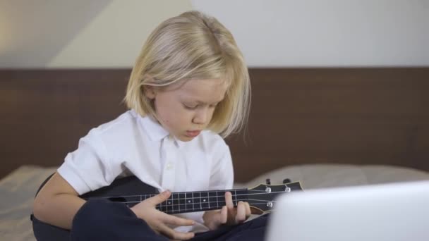 Jarak belajar pendidikan online. Seorang anak prasekolah belajar memainkan ukulele di laptop di rumah. Tes anak laki-laki mengajarkan akord — Stok Video