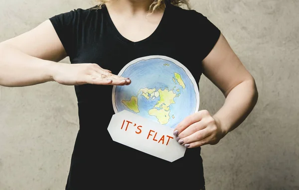 Flat Earther concept. Person who believes that Earth is flat disc. Anonymous woman holding flat Earth model in front of body with text: It`s flat. Isolated on gray background, studio shot.