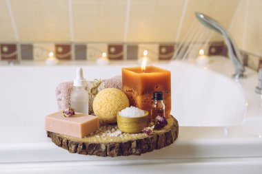 Ev yapımı spa ürünleri ahşap disk tepsisinde: sabun, banyo bombası, aroma banyosu tuzu, zaruri ve masaj yağları, mum yakma, küvet havlusu, su akışı. Rahatlatıcı konsept.
