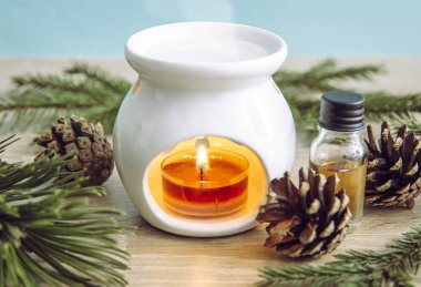 Beyaz seramik mum aromalı yağ lambası, çam ağacı ladini esaslı yağ şişesi ve ahşap ve mavi arka planda süsleme için ağaç dalları..