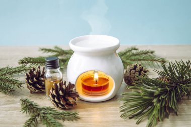 Beyaz seramik mum aromalı yağ lambası, çam ağacı ladini esaslı yağ şişesi ve ahşap ve mavi arka planda süsleme için ağaç dalları..