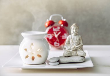 Yoğun yaşam ve sağlıklı akıl sağlığı arasında nasıl denge kurulacağını. Oturup meditasyon yapmak için ara verdim. Arka planda aroma lambası ve saati olan Buda figürü..