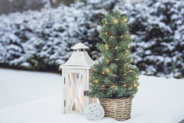 Beyaz mumlu beyaz ahşap fener ve mikro ışıklı küçük Noel ağacı karda kahverengi rattan çiçek kabı, arka planda karlı köknar ağaçları, kışın açık havada. Boşluğu kopyala.