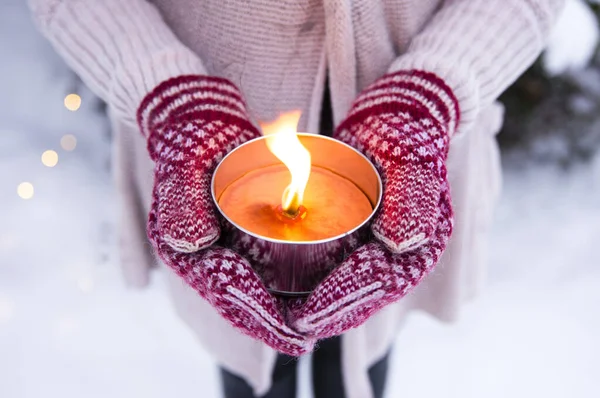 Selective focus on woman wearing knitted mittens and sweater holding a lit outdoor candle(also known as pitch torch or a garden candle), outdoors in winter, snow and bokeh lights on background.
