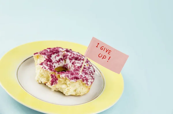 Half eaten bites taken from donut with white and pink frosting on yellow plate with surrendering flag text: I give up, on blue background. Studio conceptual shot of weight loss fail.