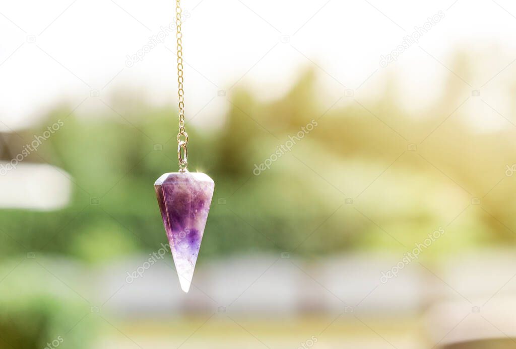 Purple natural Amethyst crystal quartz pendulum hanging on chain on green outdoors nature background.