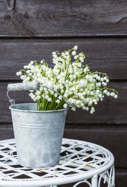Beyaz kesim çiçek buketi Vadi Zambağı (Convallaria majalis) çinko bir kova şeklinde vazoda, beyaz süslü bir çiçek standında, kahverengi ahşap arka planda.