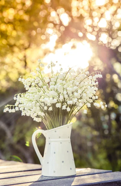 Bouquet of white flowers Lily of the valley (Convallaria majalis) also called: May bells, Our Lady's tears and Mary's tears in a white dotted jug shaped vase, outdoors on a table, trees on background.