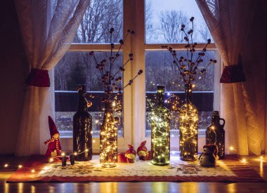 Pencerenin arkasında, içi mikro ışıklarla dolu eski bira şişeleri ve şarap şişeleriyle şirin ve sıcak bir Noel süslemesi var. Kırmızı yapay böğürtlen için vazo.