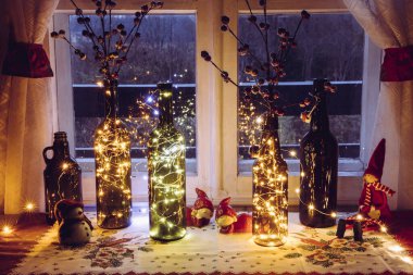 Pencerenin arkasında, içi mikro ışıklarla dolu eski bira şişeleri ve şarap şişeleriyle şirin ve sıcak bir Noel süslemesi var. Kırmızı yapay böğürtlen için vazo.