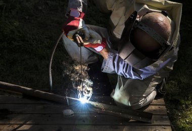 Kaynak makinesi çubuğu kullanan bir adam erimiş demir metali kesiyor açık havada, koruyucu maske ve eldiven takıyor, kıvılcımlar uçuşuyor.