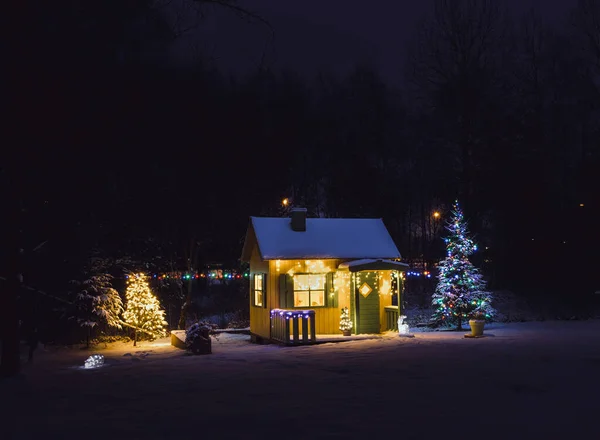 Wooden painted yellow private children`s play house in home garden, decorated with Christmas LED string lights outdoors in cold winder night. Decorated Christmas fir tree.