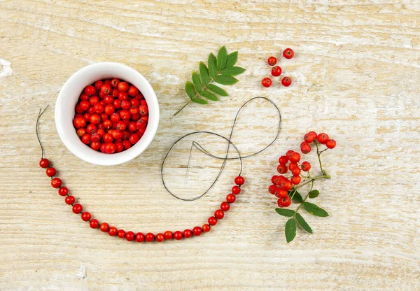 Making protective rowan berry charm necklace, it is believed that rowan tree berries have magical properties. Old folklore concept. Light natural wooden background.