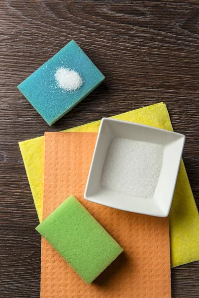 Natural organic citric acid cleaning products concept. White Citric acid powder in white square bowl and on blue, green, orange and yellow washing sponge and textile cloth on brown wooden background.