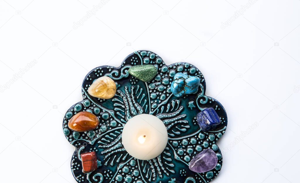 Top view of all 7 chakra colors semi precious crystal stones on blue and white ornament background with relaxing candle burning in center. Lot of copy space for text.