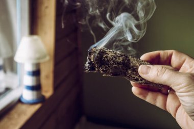 Elinde kurutulmuş adaçayı tomarı olan bir kadın sigara içiyor. Negatif enerjiyi temizlediğine ve evdeki yaşam alanlarını arındırdığına inanılıyor..