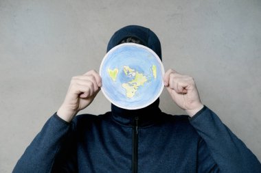 Düz Dünyalı kavramı. Dünya 'nın düz disk olduğuna inanan kişi. İsimsiz kapüşonlu adam düz Dünya modelini suratının önünde tutuyor, kopyalama uzayı. Gri arka planda, stüdyo çekimlerinde izole edilmiş..
