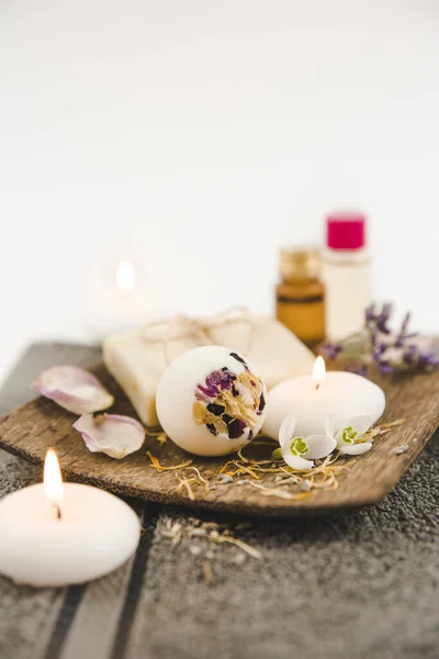 Various spa bath products on brown wooden tray creamy bath bomb, natural soap bar with cotton string, dried flower petals, spa candles lit, essential bath and massage oils in background. Copy space.