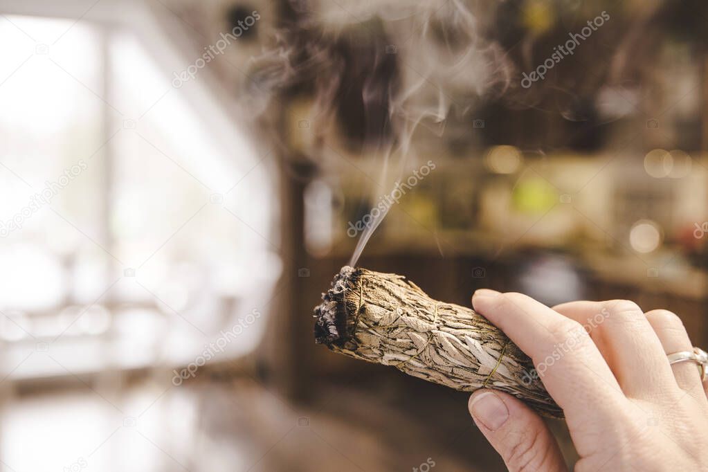 Woman hand holding herb bundle of dried sage smudge stick smoking. It is believed to cleanse negative energy and purify living spaces at home in rooms.