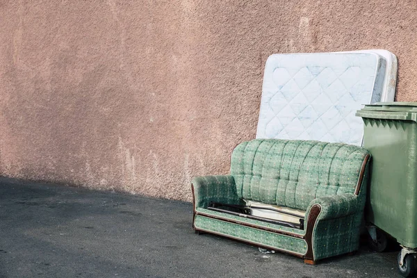 Old sofa and mattress thrown away outdoors by the house building and by the garbage can. A lot of copy space.