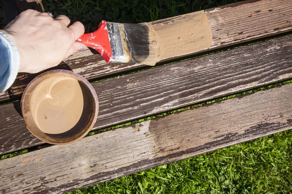 Close up of hand staining wooden furniture with brush and brown protective wood stain, repainting old terrace furniture in spring. Refreshing, giving new look concept.