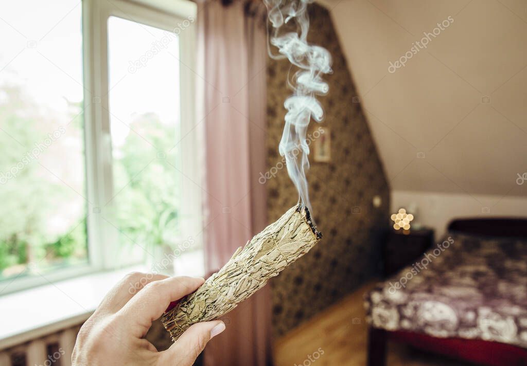 Woman hand holding herb bundle of dried sage smudge stick smoking. It is believed to cleanse negative energy and purify living spaces at home in rooms.