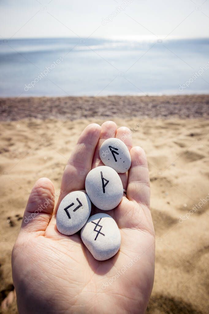 Hand holding norse rune stones with black symbols for fortune telling, beautiful outdoor sunny sandy beach with sea on background.