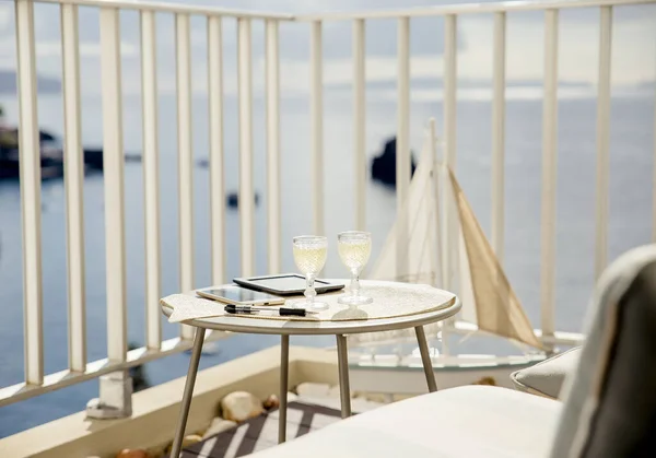 Working and traveling, busy luxurious lifestyle concept background. Shallow depth of field. Focus on white wine glasses on table, next to it are e book, cell phone and pencil. Ocean view from balcony.