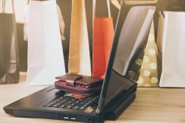 Buying from online. Virtual shopping concept. Laptop computer on wooden home living room table. Buy comfortably from home. Red wallet and credit cards paper shopping bags on the background.