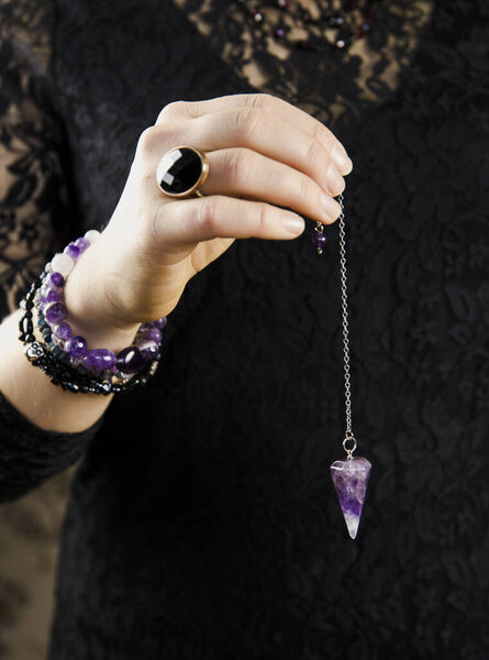 Close up of woman wearing black gothic clothing, hand holding and using amethyst crystal pendulum on silver chain fortune telling pendulum in hand. Mystical psychic dark witch concept. 