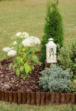 Round flower bed covered by pine bark mulch and surrounded by brown weatherproofed wood roll boarder palisade on bed are multiple different plants outdoors in summer, lantern on wooden base. clipart