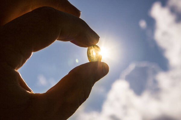Vitamin D keeps you healthy while lack of sun. Fingers holding yellow soft shell D-vitamin capsule against sun and blue sky on sunny day. Cure concept.