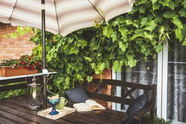 Dekorasyon olarak yemyeşil üzümleri olan şirin özel bir balkon ve güneşli yaz günlerinde güneşli bir plaj şemsiyesi. Açılmış kitap, modern metal ampul fener ve bahçe mobilyalarının üzerindeki bardak..