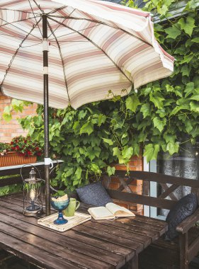 Dekorasyon olarak yemyeşil üzümleri olan şirin özel bir balkon ve güneşli yaz günlerinde güneşli bir plaj şemsiyesi. Açılmış kitap, modern metal ampul fener ve bahçe mobilyalarının üzerindeki bardak..