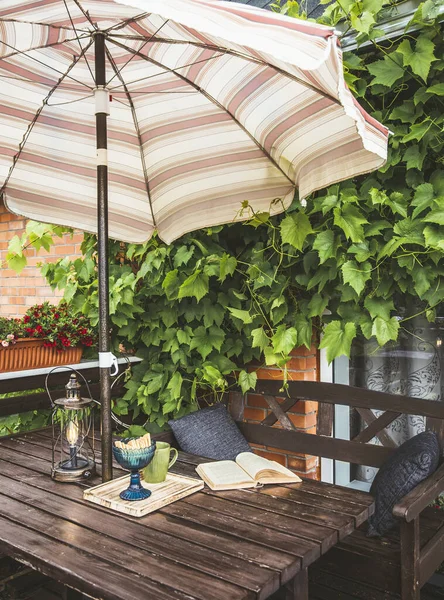 Dekorasyon olarak yemyeşil üzümleri olan şirin özel bir balkon ve güneşli yaz günlerinde güneşli bir plaj şemsiyesi. Açılmış kitap, modern metal ampul fener ve bahçe mobilyalarının üzerindeki bardak..