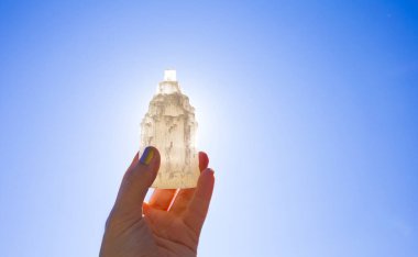 Güneş ve mavi gökyüzüne karşı doğal yontulmuş Selenit kulesi taşıyan kişi taşın iyileştirici ve arındırıcı özellikleri vardır. Ayrıca diğer mineral kristal taşlarını şarj etmek için kullanılır..