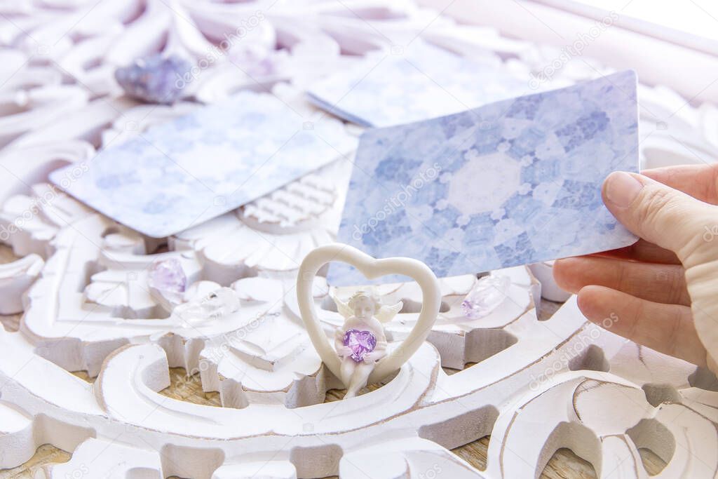 Deck with divination homemade Angel cards on bright white table, surrounded with semi precious stones crystals and candles. Selective focus on cute angel figurine. Person choosing a card.